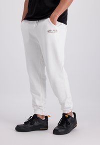 Witte sweatpants met elastische boorden en zakken, voorzien van een bruin "Alpha Industries" logo. Draag ze met zwarte Nike sneakers.
