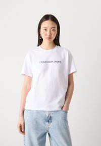 Balta medvilnės marškinėliai su trumpomis rankovėmis ir apvaliu kaklu, su juodu tekstu "Calvin Klein Jeans" per krūtinę. Atsitiktinė forma.