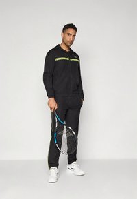 Μαύρη αθλητική φόρμα με πράσινες λεπτομέρειες και λογότυπα. Το μοντέλο κρατάει μια ρακέτα τένις. Άσπρα αθλητικά παπούτσια ολοκληρώνουν το αθλητικό σύνολο.