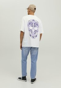 Camisa blanca de manga corta con un gráfico de calavera floral morada en la parte de atrás, combinada con jeans azul claro y zapatillas negras.