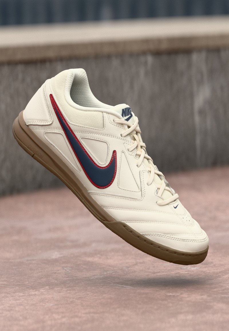 Buty sportowe Nike mają kremową skórzaną cholewkę z niebieskim i czerwonym logo, teksturowane dodatki oraz brązową gumową podeszwę. Zapięcie na sznurowadła.
