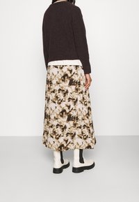 Brauner, flauschiger Pullover über einem weißen Oberteil, kombiniert mit einem langen, gemusterten Rock in Beige- und Brauntönen. Weiße Knöchelstiefel mit schwarzen Akzenten.