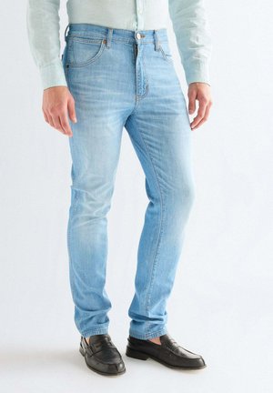 Mann trägt hellblaue Slim-Fit-Jeans, schwarze Lederloafers und ein helles, geknöpftes Hemd und steht vor schlichtem Hintergrund.