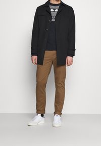 Zwarte jas met knopen, marineblauwe trui met een wit patroon, tan kleurige broek, en witte sneakers. Glad materiaal, gestructureerde pasvorm.