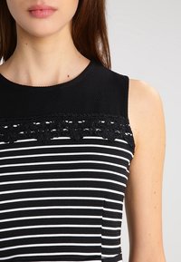 Kvinna som bär en ärmlös svart och vit randig topp med en texturerad svart övre del och spetsdetalj över bröstet.