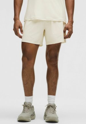 Mand iført cremefarvede atletikshorts, hvide sokker og beige løbesko, står mod en ensfarvet baggrund.