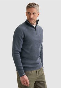 Homme aux cheveux gris portant un pull bleu côtelé à demi-fermeture éclair et un pantalon vert olive, regardant sur le côté sur un fond uni.