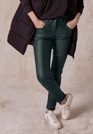 Dunkelgrüne, schmal geschnittene Hose mit glänzender Oberfläche, ausgestattet mit Vordertaschen und kombiniert mit weißen Sneakers und einer dunklen Weste.