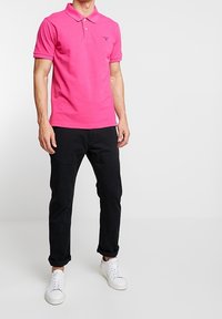 Polo shirt rosa con colletto e maniche corte, abbinato a pantaloni neri e sneakers bianche. La maglietta presenta un delicato dettaglio del logo.