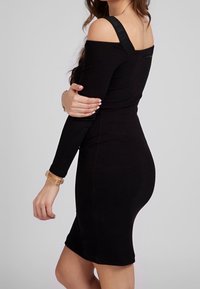 Robe noire à manches dénudées avec un design ajusté, en tissu doux et dotée d'une bretelle avec logo. La robe arrive juste au-dessus du genou.