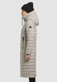 Cappotto imbottito lungo di colore grigio chiaro con cuciture verticali, cappuccio regolabile e bottoni neri di accentatura. Tasca laterale con zip visibile.