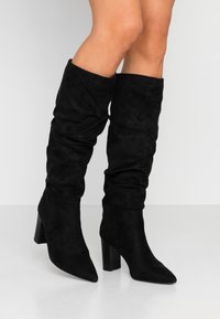Bottes noires montant jusqu'aux genoux en daim doux avec un design froncé, bout pointu et talon bloc épais. Texture lisse et apparence élégante.