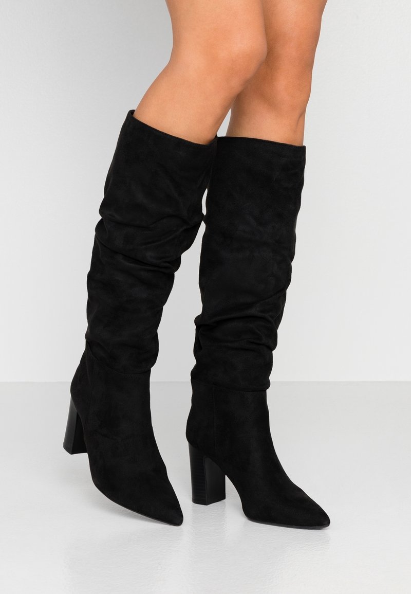 Bottes noires montant jusqu'aux genoux en daim doux avec un design froncé, bout pointu et talon bloc épais. Texture lisse et apparence élégante.