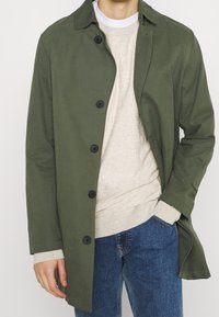 Olivgrüner, leichter Mantel mit großen schwarzen Knöpfen, Fronttaschen und Kragen, über einem beigefarbenen Pullover und blauen Jeans getragen.