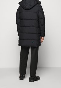 Sort pufferjakke med hætte, der har et quiltet design og en grøn detalje i kanten. Båret med mørke bukser og sorte sko.