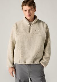 Μπεζ φλις πουλόβερ με φερμουάρ μέχρι τη μέση μπροστά, υφή επιφάνειας και στρογγυλή γιακά. Διαθέτει μικρό λογότυπο στο στήθος. Κανονική εφαρμογή.