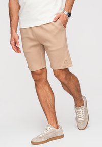 Homme portant un short beige et des baskets blanches, une main dans la poche, montre-bracelet visible au poignet gauche, debout sur un fond clair.