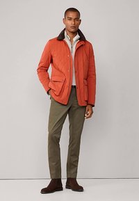 Prešívaná oranžová bunda s hnedým golierom, dvoma prednými vreckami, nosená cez svetlú košeľu a olivové nohavice, doplnená hnedými čižmami.