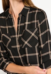 Žena v černé košili na knoflíky s béžovým kostkovaným vzorem a dvěma náprsními kapsami, rukávy vyhrnuté, ruka držící ruku u lokte.