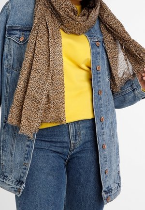 Personne portant un haut jaune, une veste en jean bleue et un jean, tenant une écharpe à imprimé léopard autour du cou d'une main.