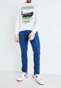 White long-sleeve shirt with black and green graphic, paired with blue slim-fit pants and white sneakers with red and blue accents.