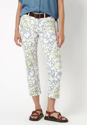 Frau trägt weiße verkürzte Hose mit blauem Blumenmuster, braunem Gürtel, blauem Oberteil und braunen Sandalen, steht vor einem einfarbigen Hintergrund.