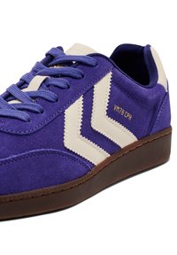Fioletowy sneaker z zamszu z białymi zygzakowymi akcentami i złotym napisem "VM78 CPH." Brązowa gumowa podeszwa z fakturą. Płaskie sznurowadła.