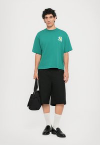 Green short-sleeve t-shirt with graphic, black knee-length shorts, white socks, black loafers, and a black bag. Plain background.