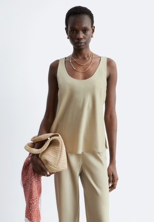 Femme portant un haut sans manches beige et un pantalon, tenant un sac beige tissé avec un foulard rouge à motifs posé dessus.