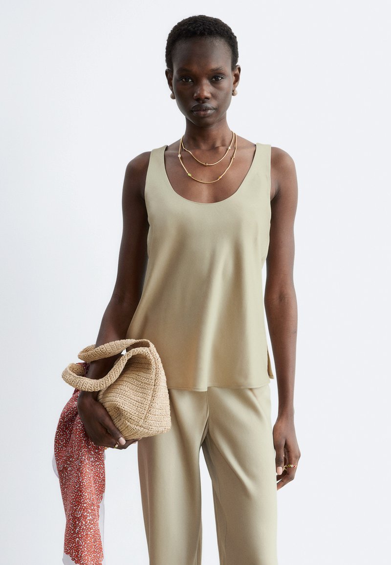 Femme portant un haut sans manches beige et un pantalon, tenant un sac beige tissé avec un foulard rouge à motifs posé dessus.