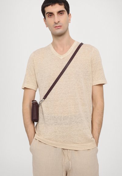 Beige V-Ausschnitt-T-Shirt aus leichtem Stoff, kombiniert mit beigen Jogginghosen mit Kordelzug. Ein dunkelbrauner Schulterriemen mit einer kleinen Tasche hängt diagonal.