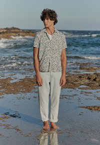 Joven con camisa de manga corta estampada y pantalones claros remangados, de pie descalzo en una orilla rocosa y húmeda con olas al fondo.