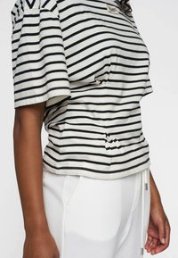 T-shirt a righe beige con strisce orizzontali nere, dotata di maniche corte, dettaglio arricciato sul lato e un'etichetta sul colletto.