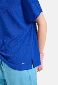 Top de algodón azul con un patrón floral texturizado, mangas cortas y aberturas laterales. Combinado con pantalones de mezclilla azul claro.