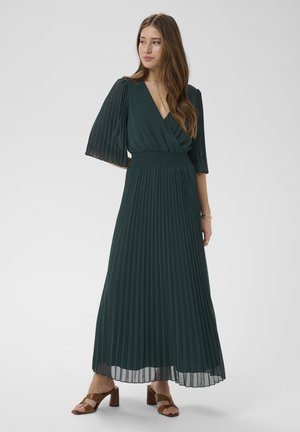 Femme portant une robe maxi plissée vert foncé avec des manches trois-quarts, une taille cintrée et des sandales marron à talons, debout devant un fond uni.
