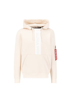 Cream-colored hoodie with front kangaroo pocket, vertical "Alpha Industries" text, drawstrings, and a small zip pocket with red "Remove Before Flight" tag on sleeve.