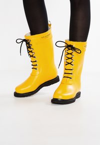 Bottes de pluie en caoutchouc jaunes avec des lacets noirs, une semelle texturée et des œillets en argent. Logo visible sur le côté. Fini lisse et brillant.