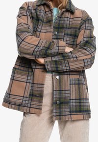 Chaqueta de franela a cuadros en tonos marrones, verdes y azules. Presenta un cuello con solapas, botones y un único bolsillo en el pecho. Tejido texturizado.