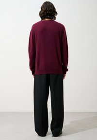 Burgundy neulepaita, jossa on pyöreä kaula-aukko, tippuvat olkapäät ja kirjailut hihansuut, yhdistettynä mustiin leveisiin housuihin.