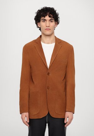 Blazer in camoscio marrone con due bottoni, colletto a revers, due tasche frontali e una finitura testurizzata. Indossato sopra una maglietta bianca.