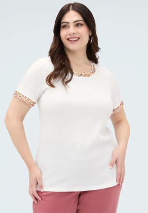 Uśmiechnięta kobieta o długich ciemnych włosach nosi biały teksturowany top z krótkim rękawem z wielokolorową koralikową lamówką, zestawiony z brązowofioletowymi spodniami.