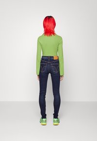 Grön långärmad topp, mörka denimjeans med en beige läderlapp bak, och ljusgröna sneakers med gula detaljer.