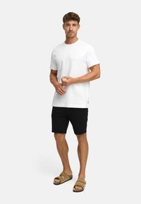 Hombre de pie que lleva una camiseta blanca simple, pantalones cortos negros y sandalias beige sobre un fondo gris claro, ajustándose las manos.