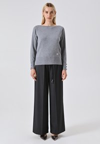 Maglione grigio lavorato a maglia con scollatura a barca, maniche lunghe e dettagli con bottoni sui polsini. Abbinato a pantaloni neri a gamba larga a vita alta.