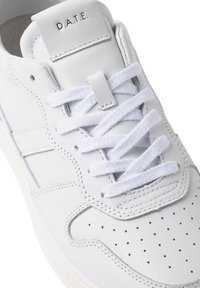 Hvide lædersneakers med perforeret tå, flade hvide snørebånd og et mærket labels på tungen. Minimalistisk design med diskrete syninger.