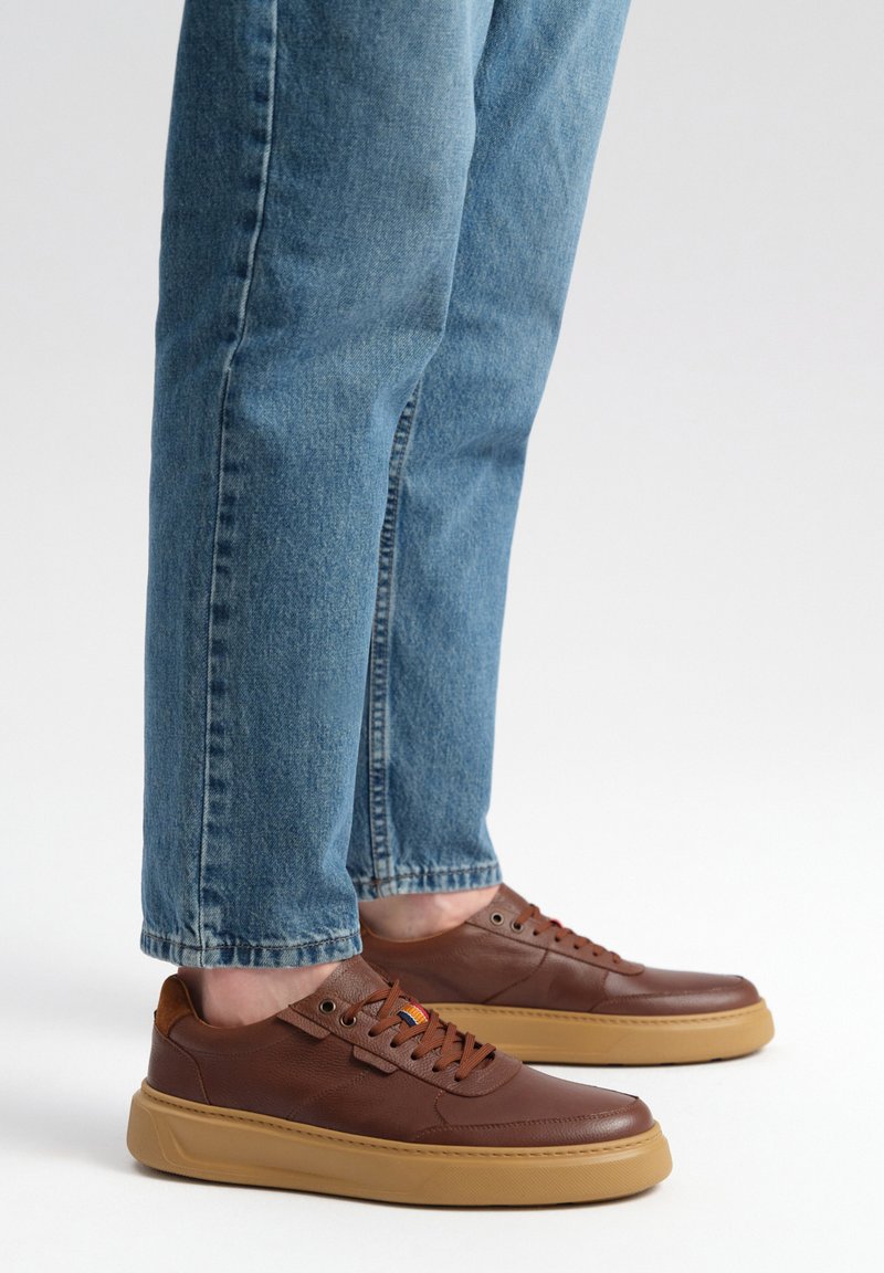 Brązowe skórzane sneakersy z teksturowanym wykończeniem, gumową podeszwą w kolorze gumy i pomarańczowymi akcentami. W zestawieniu z jasnoniebieskimi dżinsami.