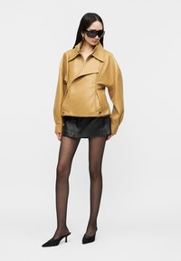 Γυναίκα που φοράει υπερμεγέθη καφέ δερμάτινο μπουφάν, μαύρη δερμάτινη μίνι φούστα, διαφανές μαύρο καλσόν, μαύρες γόβες με χαμηλό τακούνι και μεγάλα σκουρόχρωμα γυαλιά ηλίου.