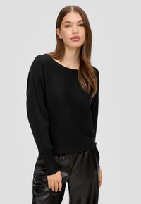 Schwarzer, gerippter Pullover mit weitem Ausschnitt und langen Ärmeln, kombiniert mit glänzenden schwarzen Kunstlederhosen. Glatte Textur insgesamt.