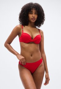 Σετ κόκκινου μπικίνι με στριφτό σχέδιο στο σουτιέν, ρυθμιζόμενους ιμάντες και διακοσμητικά μεταλλικά στοιχεία, σε συνδυασμό με ταιριαστά σλιπ χαμηλής μέσης.