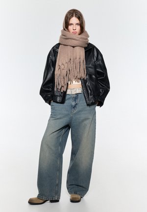 Giacca oversize in pelle nera, sciarpa oversize beige con frange, cintura bianca aderente, jeans larghi in denim chiaro e stivaletti beige alla caviglia.